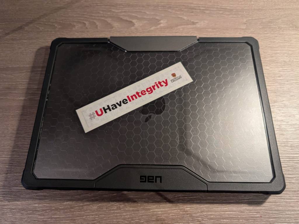 AltText: The image shows a closed laptop with a honeycomb-patterned cover on a wooden surface. On top of the laptop, there is a rectangular sticker that reads "#UHaveIntegrity" with the "integrity" part in red text. The sticker also includes a small logo for the University of Calgary.